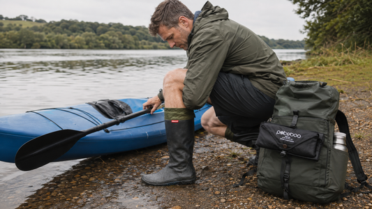 Man sat on a riverbank next to kayak wearing pokeboo TREAD packable boots