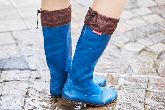 A person wearing royal blue Pokeboo  rain boots with brown waterproof cuffs, standing on wet stone pavement. A small red Pokeboo brand tag is visible on the side of the right boot.