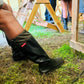 A person wearing black original Pokeboo rubber knee-high boots with a small red 'Pokeboo' brand tag, sitting outdoors on grass next to wooden structures.