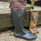 A single tall rubber rain boot by Pokeboo standing upright on a concrete surface next to a weathered wooden dock post with rope. The boot has a dark charcoal gray textured lower section and a brown fabric cuff with a small red Pokeboo brand label.