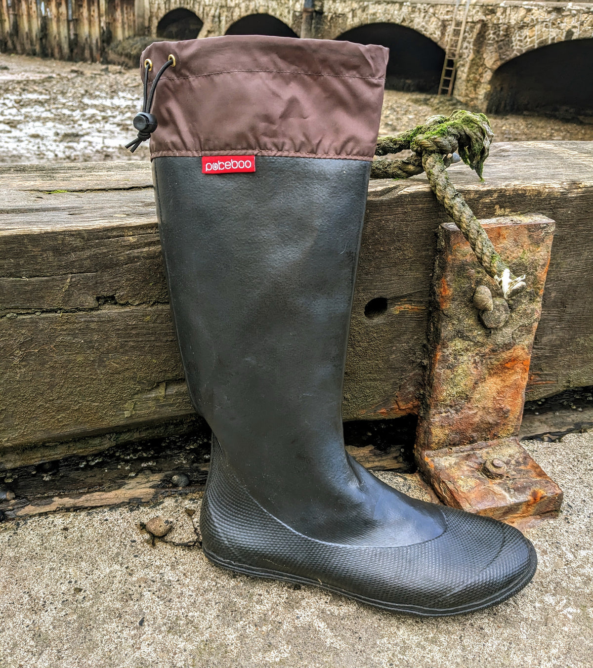 A single tall rubber rain boot by Pokeboo standing upright on a concrete surface next to a weathered wooden dock post with rope. The boot has a dark charcoal gray textured lower section and a brown fabric cuff with a small red Pokeboo brand label.