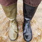 A pair of knee high Pokeboo rubber boots standing in shallow muddy water. One boot is a muted khaki green and the other is black, both showing wet, textured surfaces and mud stains.