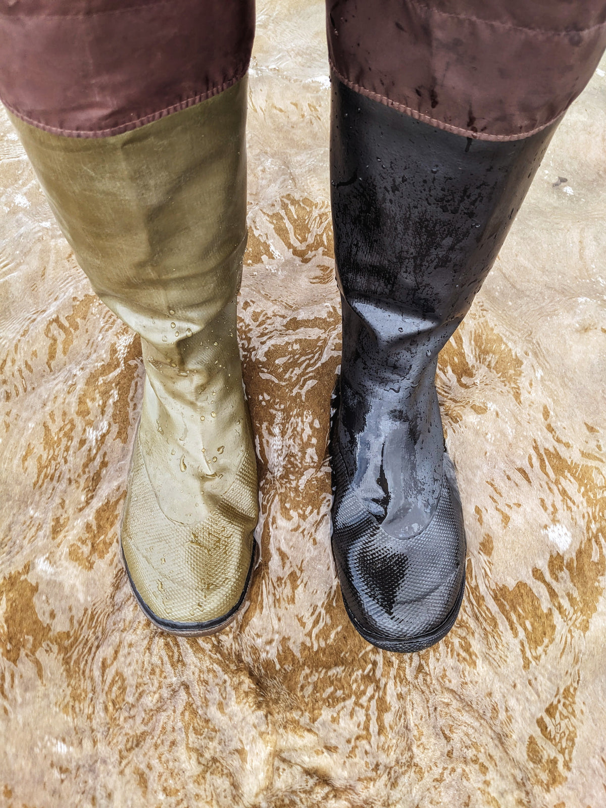 A pair of knee high Pokeboo rubber boots standing in shallow muddy water. One boot is a muted khaki green and the other is black, both showing wet, textured surfaces and mud stains.