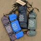 Four rolled pairs of Pokeboo waterproof boot bags in charcoal, blue, black, and khaki green, laid out on a sandy beach with seaweed.