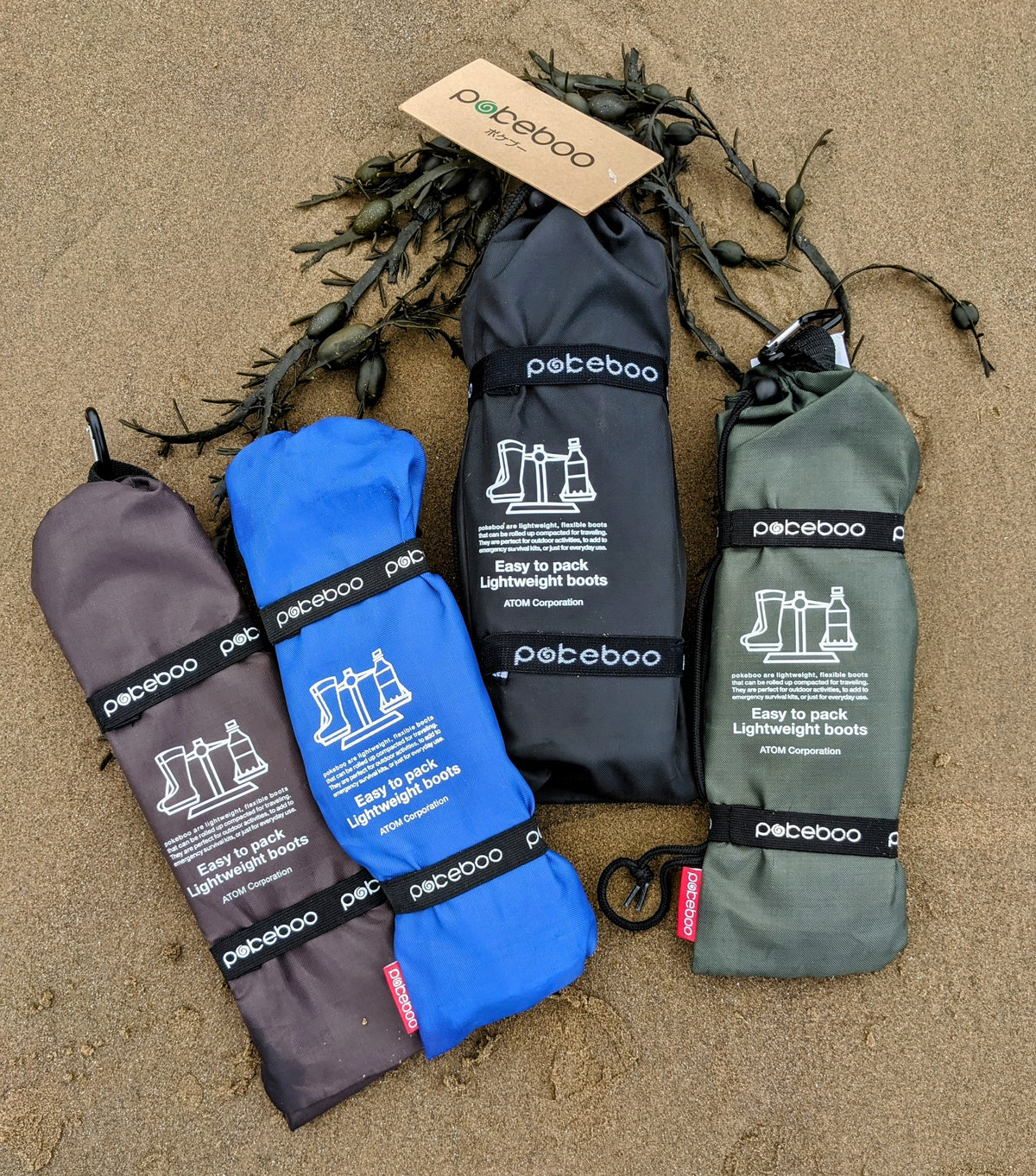 Four rolled pairs of Pokeboo waterproof boot bags in charcoal, blue, black, and khaki green, laid out on a sandy beach with seaweed.