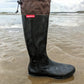 A single black original pokeboo rubber rain boot with a brown fabric cuff stands on wet sand at the beach, with ocean waves and an overcast sky in the background. A red label on the side reads 'Pokeboo'.