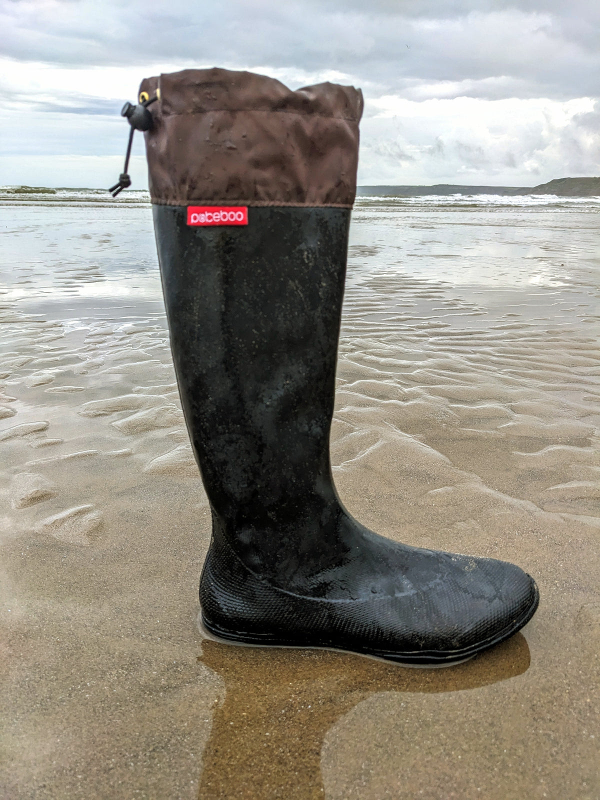 A single black original pokeboo rubber rain boot with a brown fabric cuff stands on wet sand at the beach, with ocean waves and an overcast sky in the background. A red label on the side reads 'Pokeboo'.