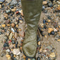 An khaki green rubber Pokeboo knee length wading boot standing in shallow muddy water on a bed of wet multicolored pebbles and stones.