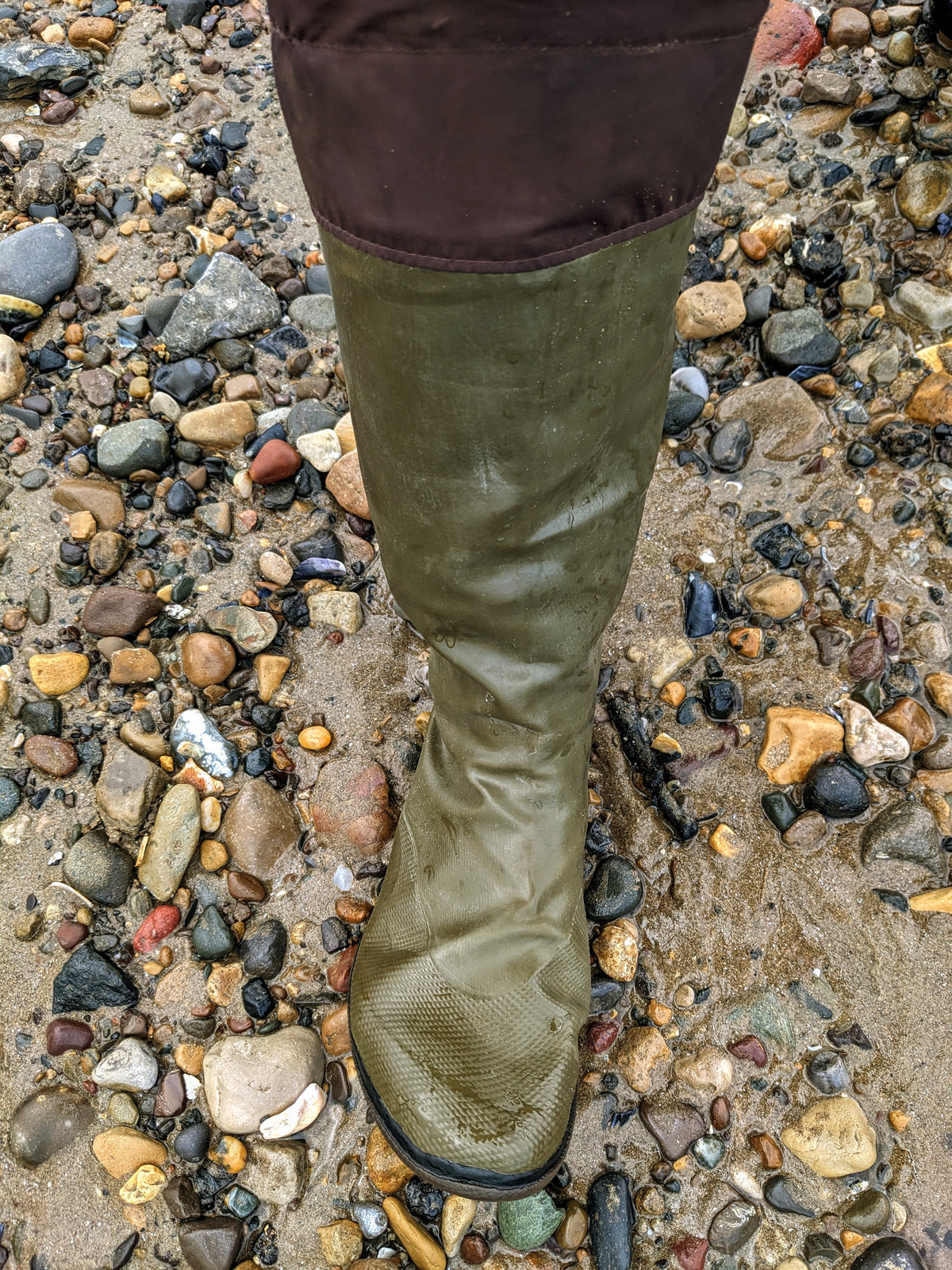 An khaki green rubber Pokeboo knee length wading boot standing in shallow muddy water on a bed of wet multicolored pebbles and stones.