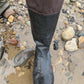 A close-up of a knee high original Pokeboo black rubber boot with a brown fabric cuff, shown standing on wet sand and rocks outdoors.