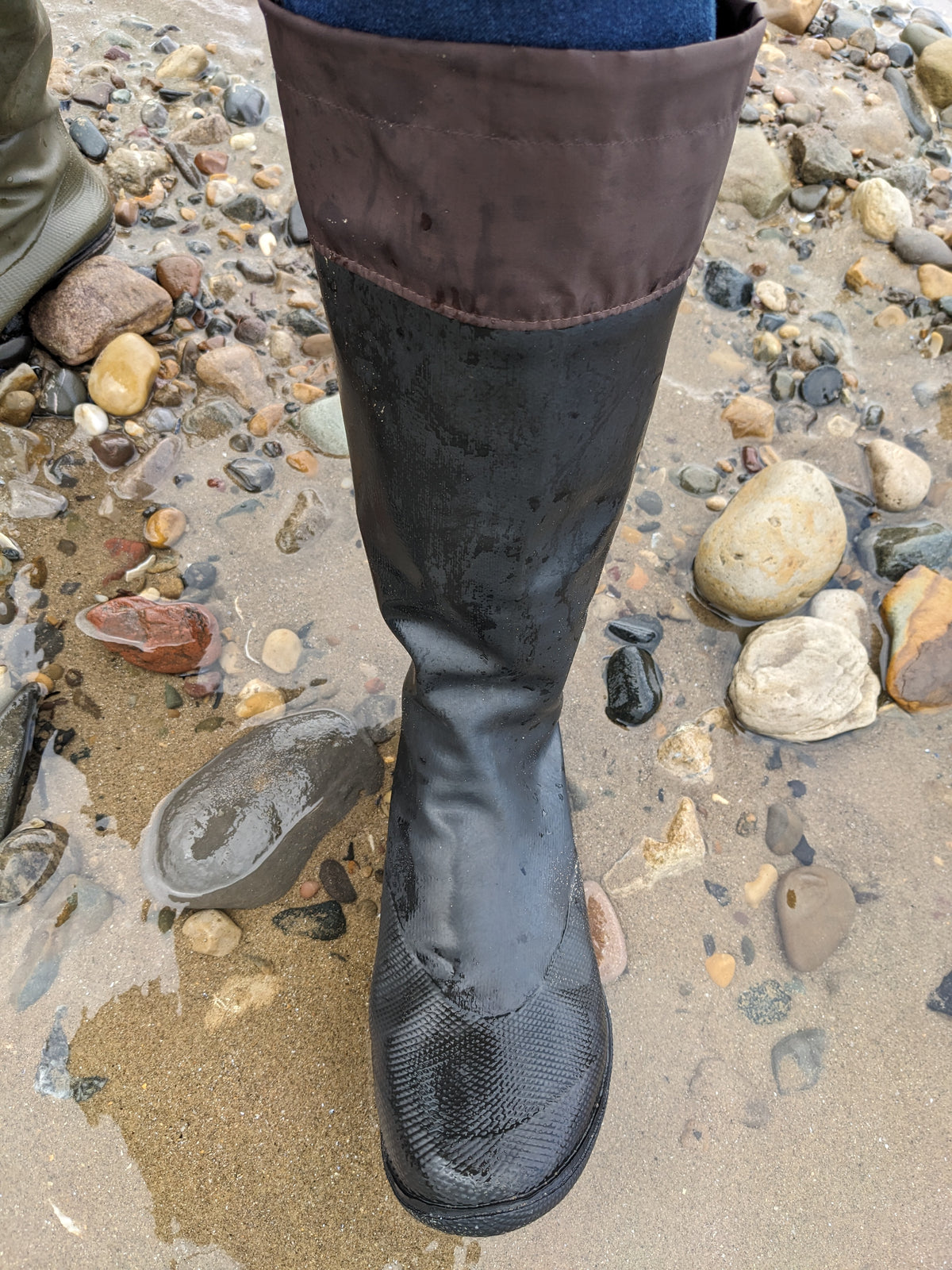 A close-up of a knee high original Pokeboo black rubber boot with a brown fabric cuff, shown standing on wet sand and rocks outdoors.