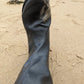 A close-up of an original Pokeboo black rubber boot standing on sandy beach ground, with footprints and sand visible around it.