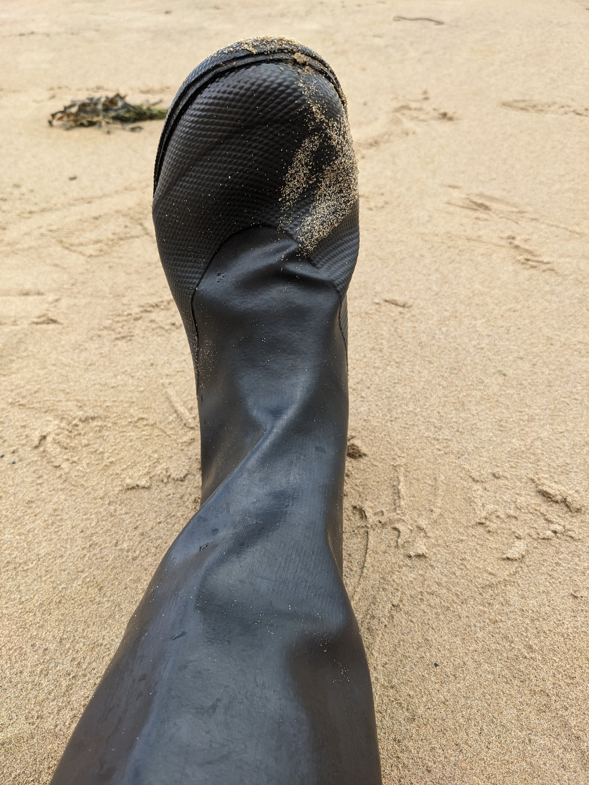A close-up of an original Pokeboo black rubber boot standing on sandy beach ground, with footprints and sand visible around it.