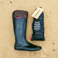 A pair of black original Pokeboos knee high waterproof boots with a brown cuff, shown next to their black carrying pouch on a sandy beach background.