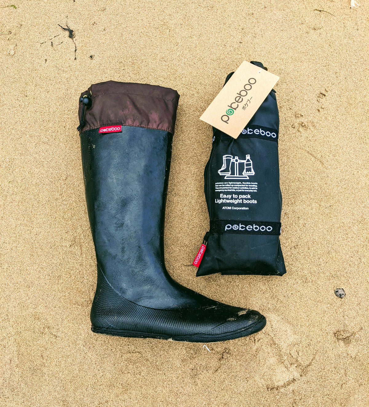 A pair of black original Pokeboos knee high waterproof boots with a brown cuff, shown next to their black carrying pouch on a sandy beach background.