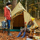 A man and a woman are camping outdoors in a wooded area with a tan tent behind them. The man wears an orange-red jacket, blue jeans, and charcoal Pokeboo rubber boots, while the woman wears a yellow beanie, a tan jacket, patterned leggings, and royal blue Pokeboo boots.
