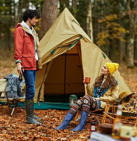 A man and a woman are camping outdoors in a wooded area with a tan tent behind them. The man wears an orange-red jacket, blue jeans, and charcoal Pokeboo rubber boots, while the woman wears a yellow beanie, a tan jacket, patterned leggings, and royal blue Pokeboo boots.