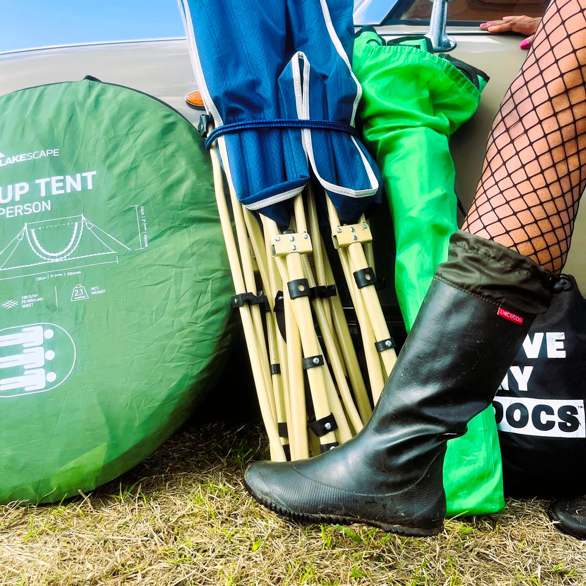 A close-up outdoor shot showing camping gear beside a car: a green round tent cover labeled 'CAMPSCAPE UP TENT PERSON,' bundled blue and green fabric items, a set of wooden folding chairs, and a person's lower leg wearing a original Pokeboo black rain boots and fishnet tights.