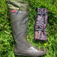 A dark olive green/charcoal Pokeboo rubber rain boot lies on green grass next to its folded brown protective cover, which has the 'pokeboo' brand name visible.