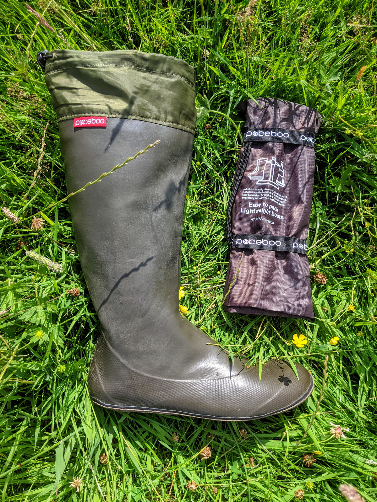 A dark olive green/charcoal Pokeboo rubber rain boot lies on green grass next to its folded brown protective cover, which has the 'pokeboo' brand name visible.