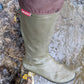 A single knee high Pokeboo rubber boot standing in shallow water. The boot is olive green with a dusty mauve-brown upper cuff and a small red Pokeboo brand tag.