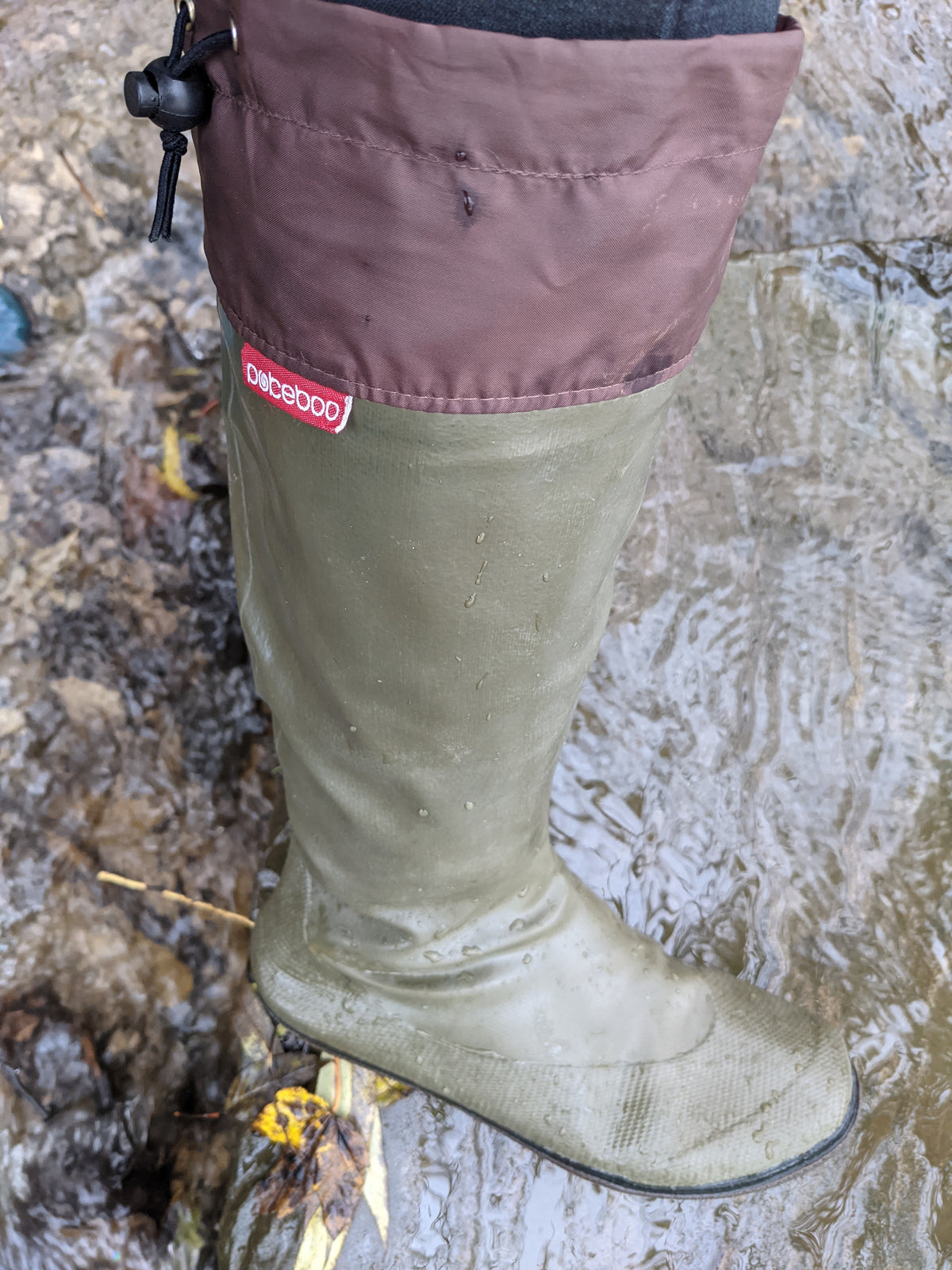 A single knee high Pokeboo rubber boot standing in shallow water. The boot is olive green with a dusty mauve-brown upper cuff and a small red Pokeboo brand tag.
