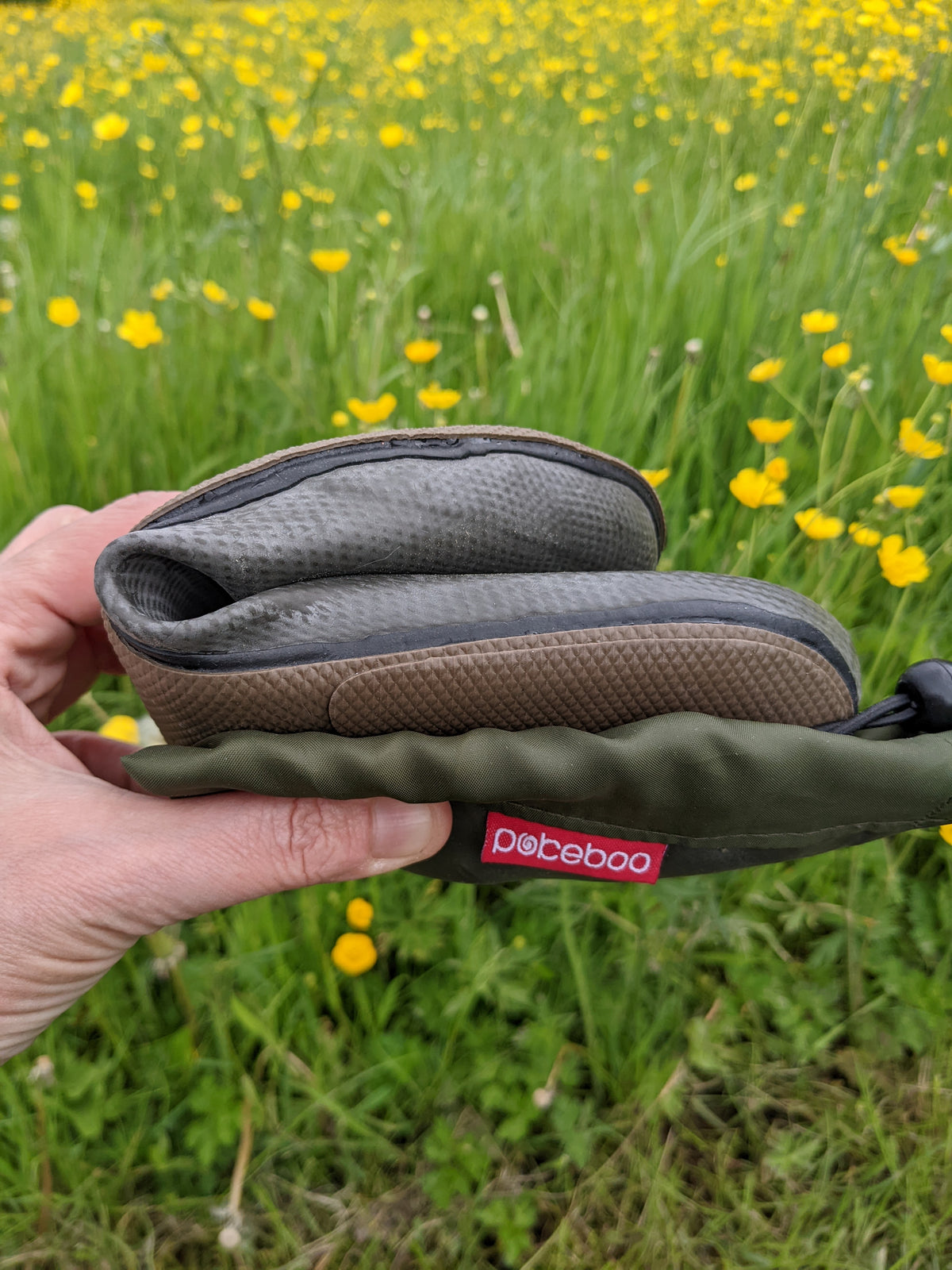 A hand holds a Pokeboo boot rolled up in their hand in front of a blurred field of yellow flowers. A red brand tag reading 'pokeboo' is visible on the pouch.