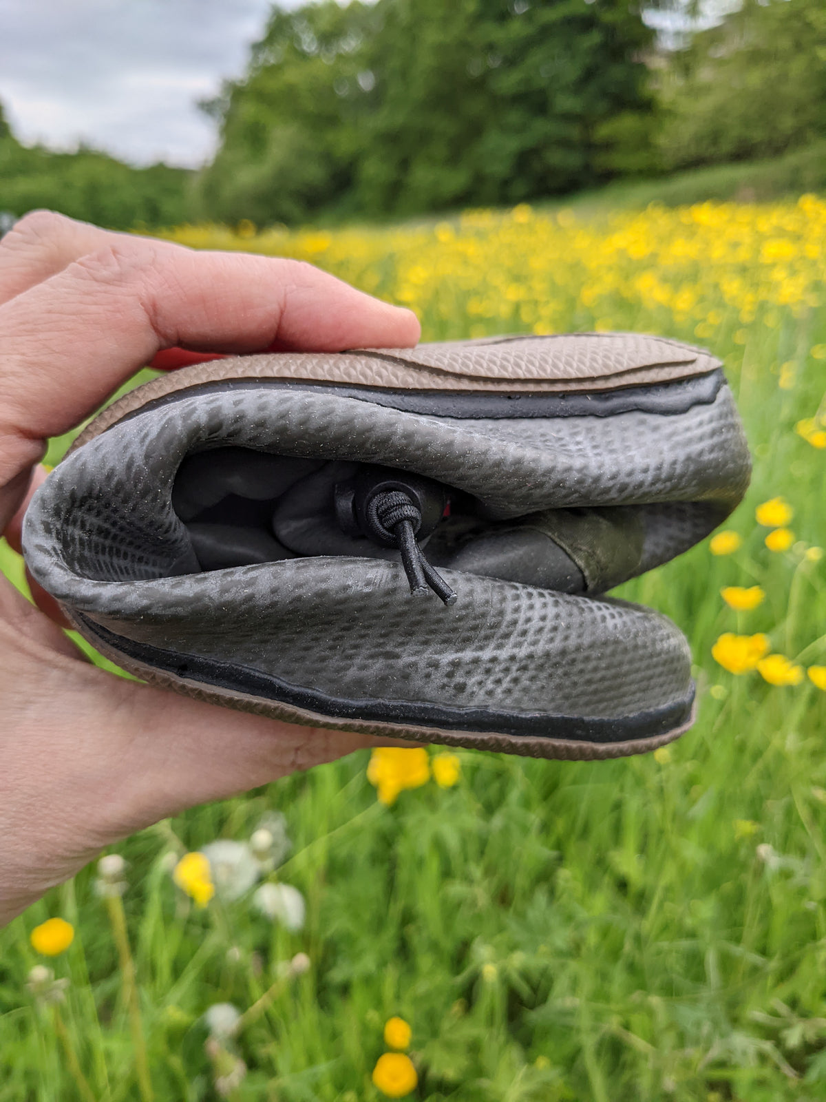 A close-up of a hand holding a rolled up Pokeboo boot in a grassy field with yellow flowers in the background.