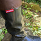 A close-up of a person's leg wearing a pair of black original Pokeboo rubber rain boots with a brown waterproof leg cover, standing outdoors on a wet log with green plants in the background. The red 'pokeboo' brand tag is visible on the side.