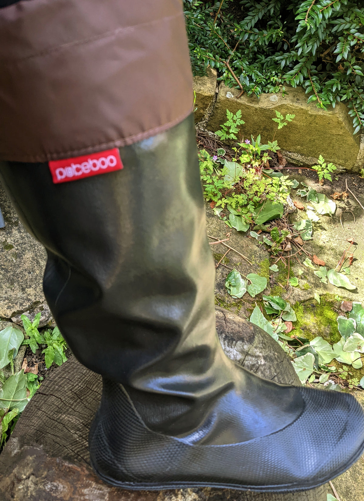 A close-up of a person's leg wearing a pair of black original Pokeboo rubber rain boots with a brown waterproof leg cover, standing outdoors on a wet log with green plants in the background. The red 'pokeboo' brand tag is visible on the side.