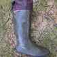 A single knee length Pokeboo rubber rain boot in khaki green with a dark brown cuff, placed on a mossy, rocky outdoor ground surface. A small red 'Pokeboo' brand tag is visible on the cuff.