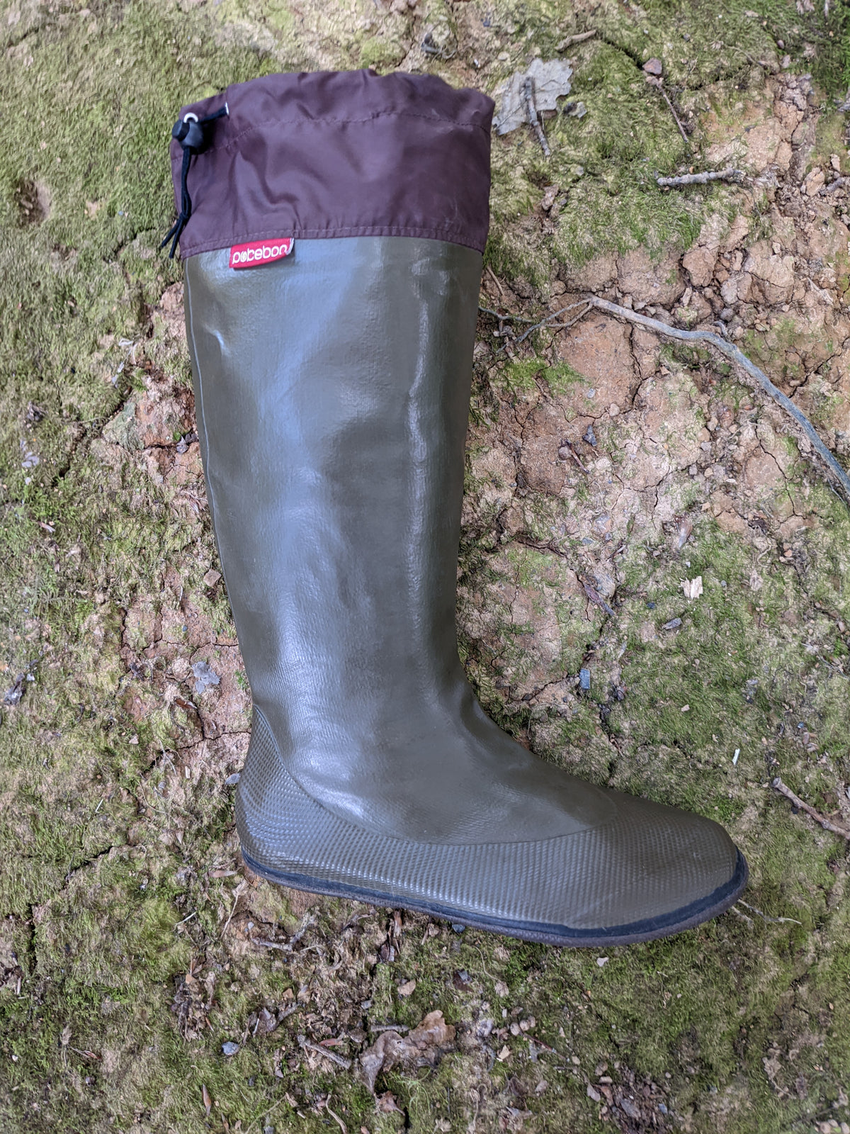 A single knee length Pokeboo rubber rain boot in khaki green with a dark brown cuff, placed on a mossy, rocky outdoor ground surface. A small red 'Pokeboo' brand tag is visible on the cuff.