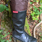 An original Pokeboo black rubber rain boot with a brown fabric cuff, shown on a mossy log in an outdoor forest setting. The red 'pokeboo' brand tag is visible on the side.