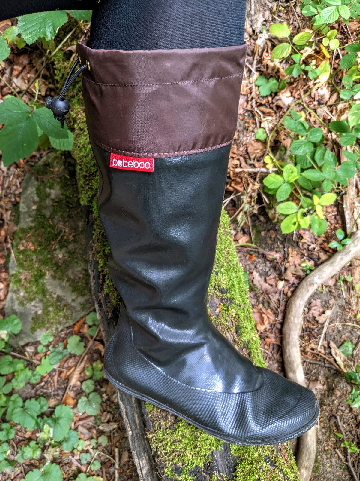 An original Pokeboo black rubber rain boot with a brown fabric cuff, shown on a mossy log in an outdoor forest setting. The red 'pokeboo' brand tag is visible on the side.