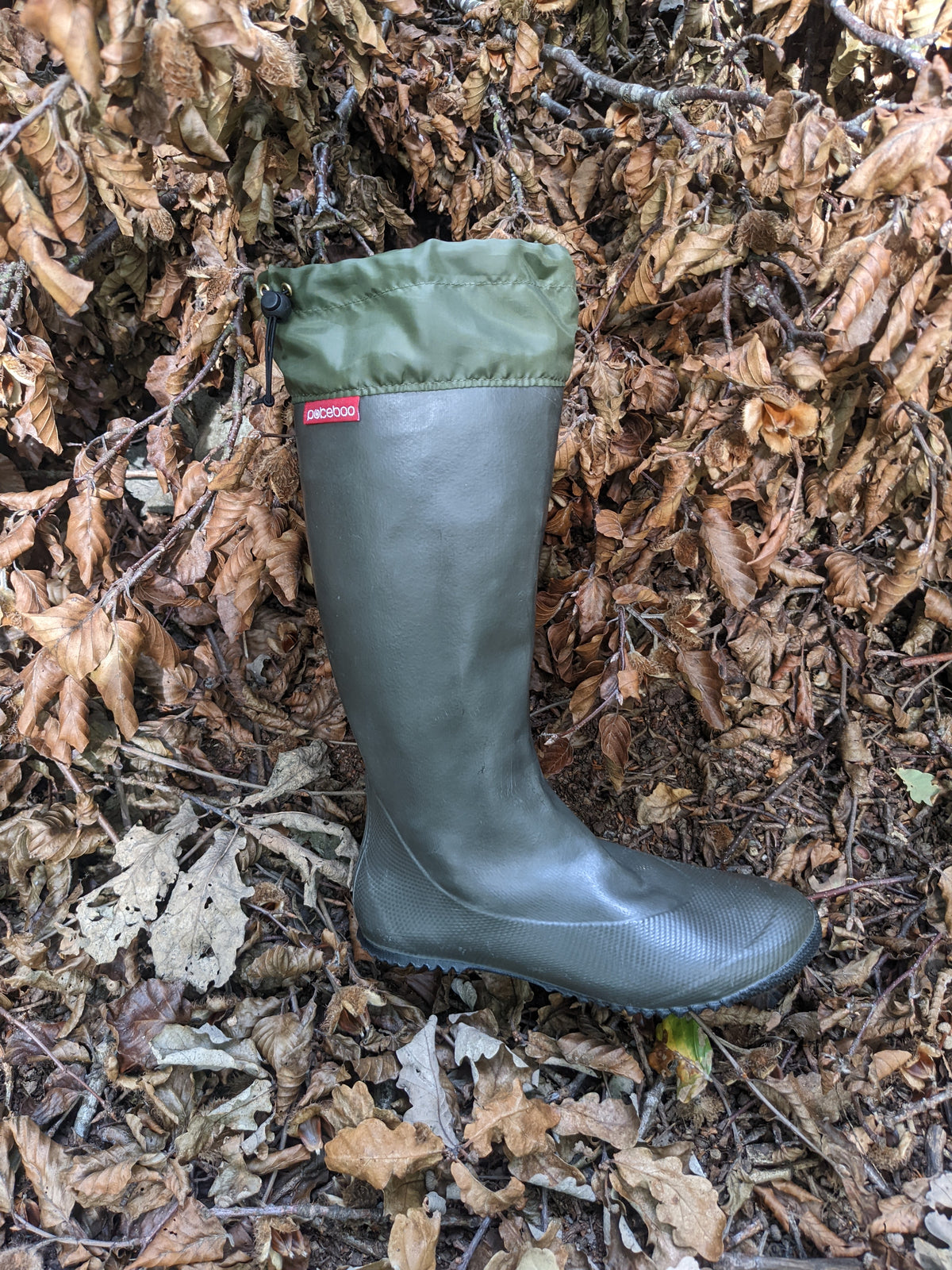 A single dark olive green/charcoal rubber rain boot with a matching green cuff, placed on a bed of dry brown leaves outdoors. A small red tag on the side of the boot shows the brand 'Pokeboo'.