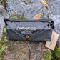 A black Pokeboo Tread bag with a brown button closure and white logo print, resting on a mossy rock next to a flowing stream. A kraft paper hangtag is attached to the side.