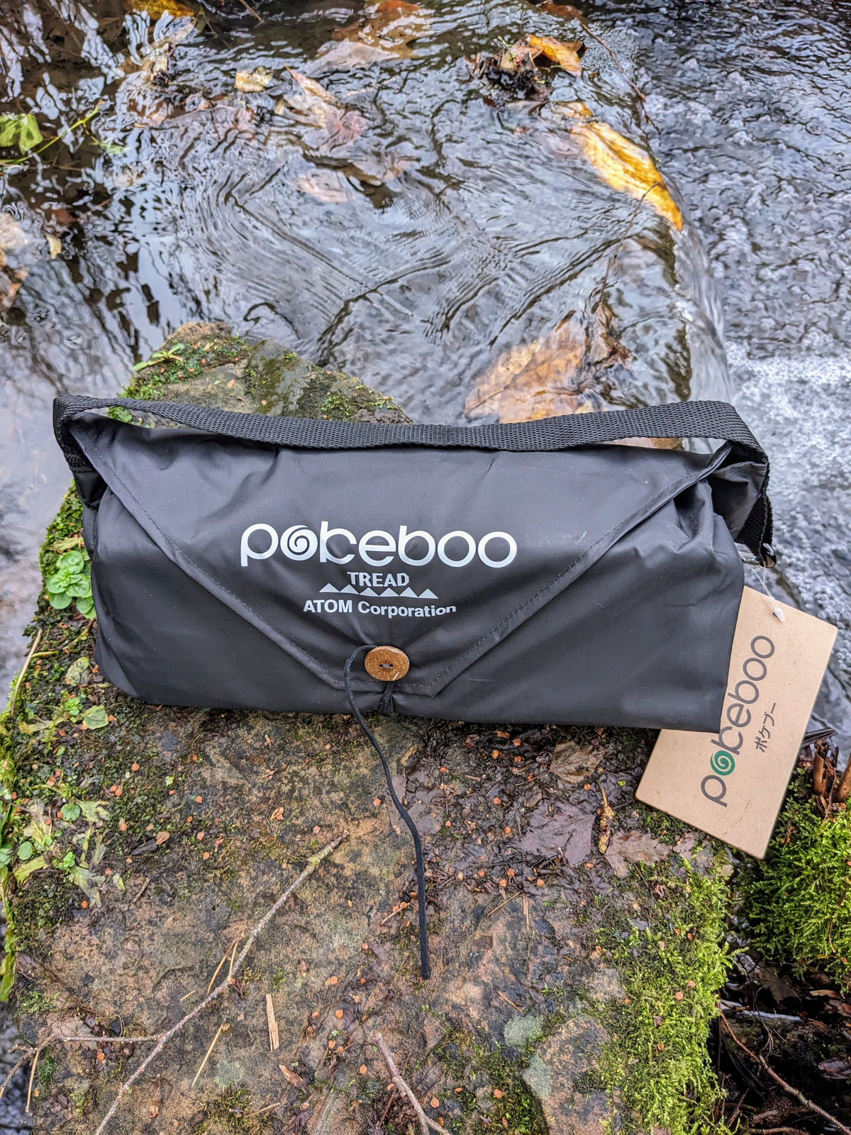 A black Pokeboo Tread bag with a brown button closure and white logo print, resting on a mossy rock next to a flowing stream. A kraft paper hangtag is attached to the side.