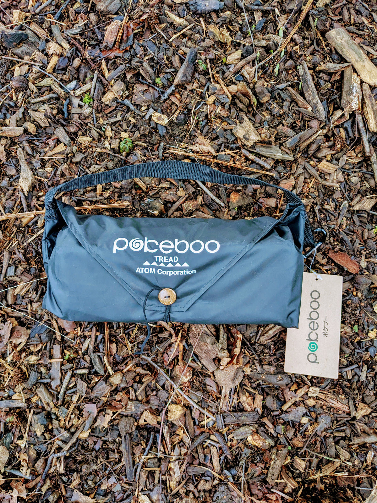 A black blue Pokeboo Tread bag laid flat on a bed of brown wood chips and leaves, with its white brand tag attached.