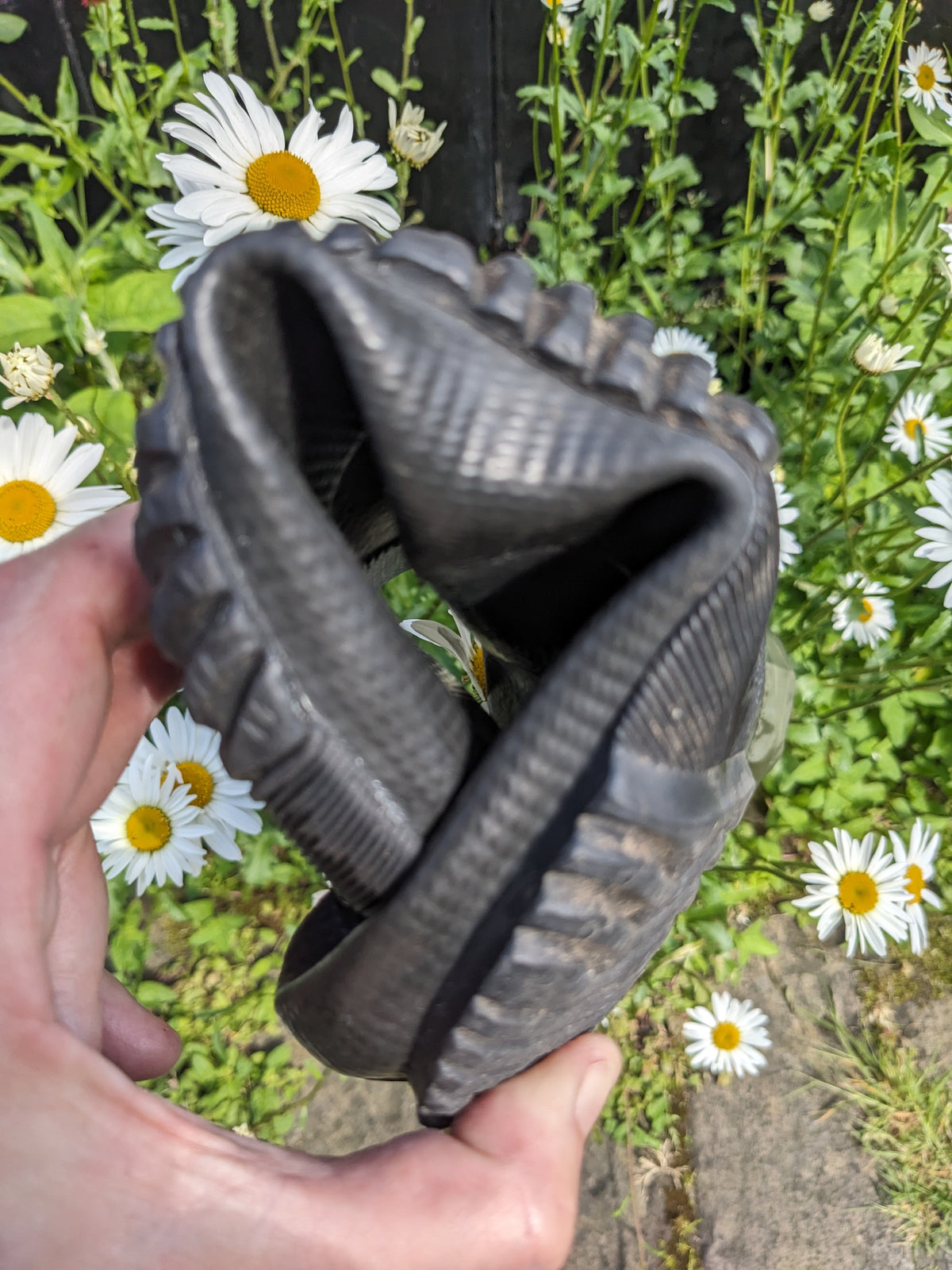 A hand holding a pair used Pokeboo TREADr ubber boots against a background of green foliage and white daisies.