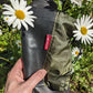 A hand holds a pair of Pokeboo TREAD rain boots outdoors among white daisies. The boots are black with an olive green hood and a small red tag that reads 'pokeboo'.