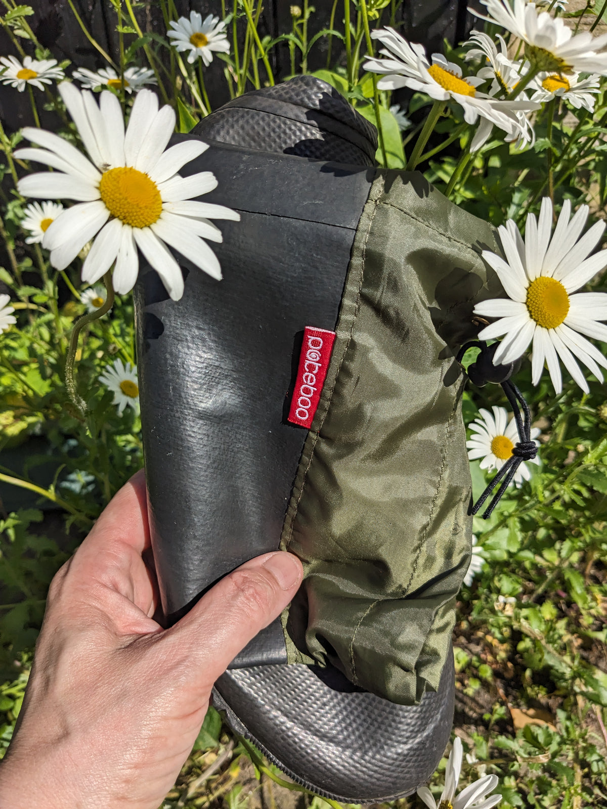 A hand holds a pair of Pokeboo TREAD rain boots outdoors among white daisies. The boots are black with an olive green hood and a small red tag that reads 'pokeboo'.