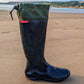 A single black Pokeboo TREAD rubber rain boot with an olive green fabric cuff, standing upright on a sandy beach with water and hills in the background. A small red tag on the side of the boot reads 'Pokeboo'.
