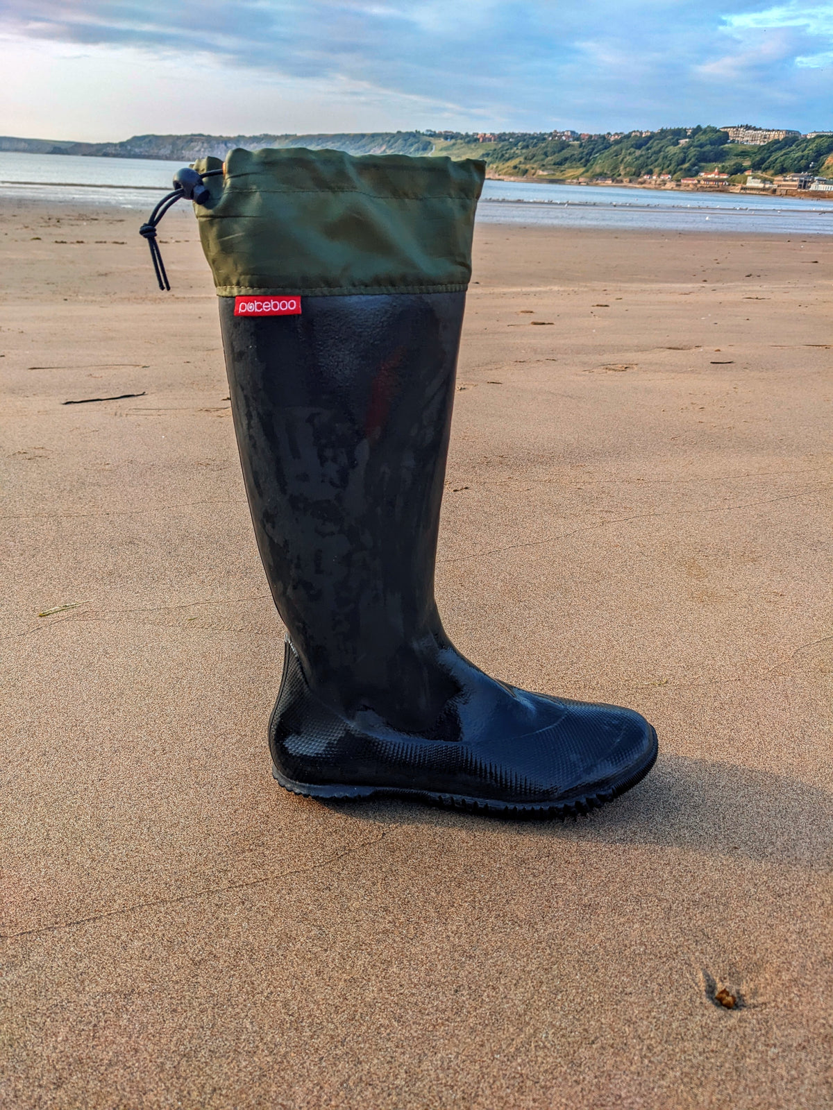A single black Pokeboo TREAD rubber rain boot with an olive green fabric cuff, standing upright on a sandy beach with water and hills in the background. A small red tag on the side of the boot reads 'Pokeboo'.