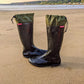 A pair of black Pokeboo TREAD rubber rain boots with olive green drawstring cuffs, placed on a sandy beach with the sea and hills in the background.