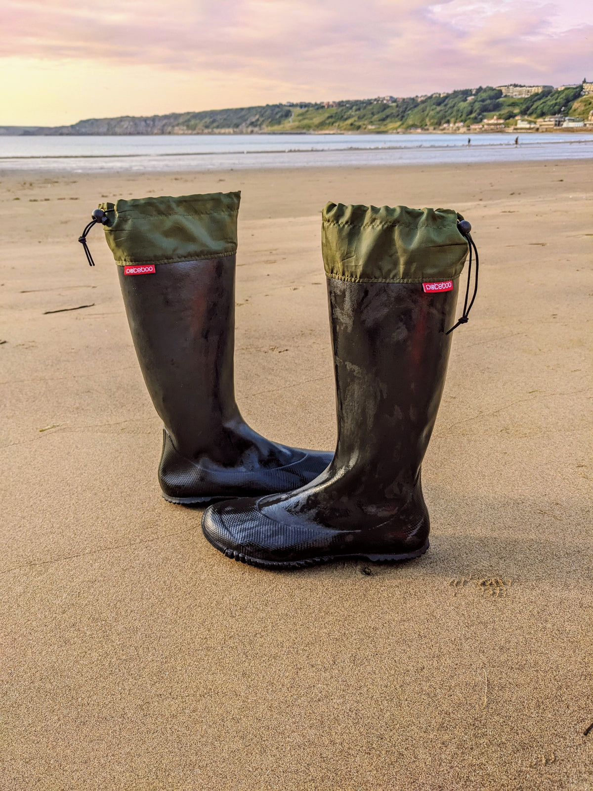 A pair of black Pokeboo TREAD rubber rain boots with olive green drawstring cuffs, placed on a sandy beach with the sea and hills in the background.