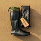 A black Pokeboo TREAD rubber rain boot with an olive green cuff sits next to its black carrying pouch on a sandy ground. The brand name 'pokeboo' is clearly visible on both the pouch and the hangtag.