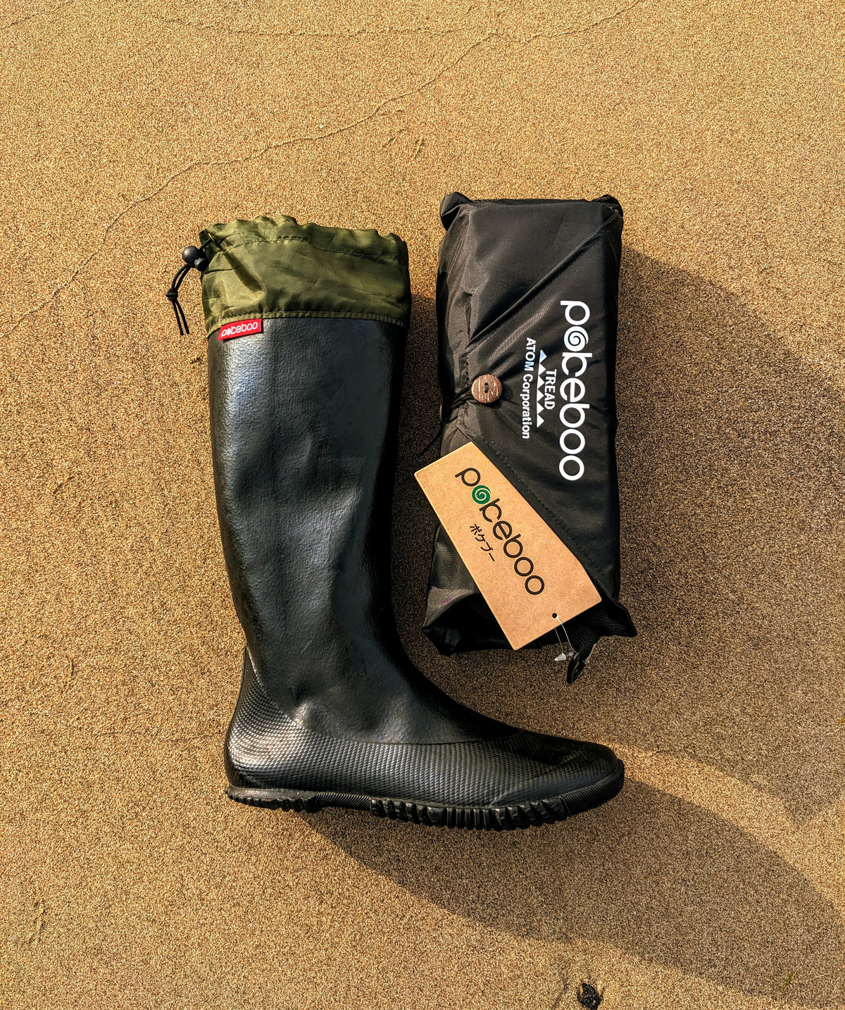 A black Pokeboo TREAD rubber rain boot with an olive green cuff sits next to its black carrying pouch on a sandy ground. The brand name 'pokeboo' is clearly visible on both the pouch and the hangtag.