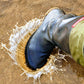 A black rubber Pokeboo TREAD rain boot with an olive green cuff is being pushed into wet sand, creating a splash of water. A small red brand tag is visible on the side of the boot.