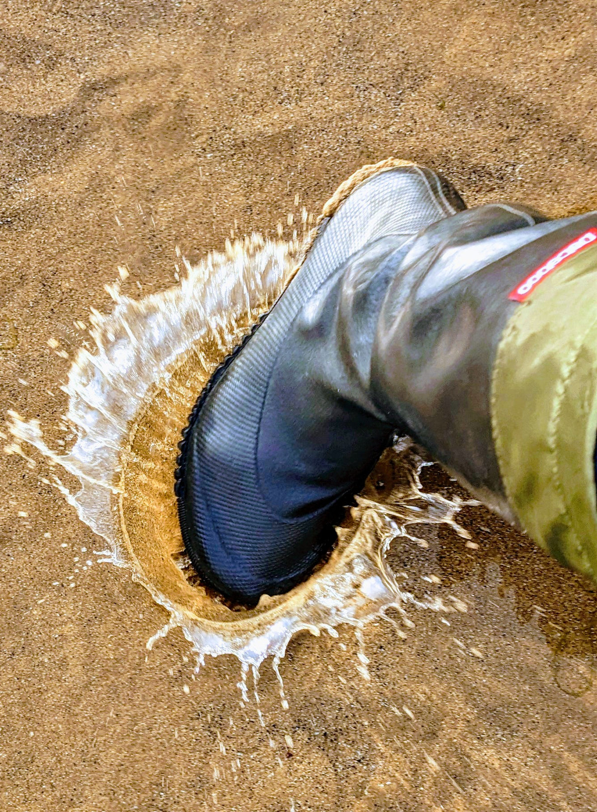 A black rubber Pokeboo TREAD rain boot with an olive green cuff is being pushed into wet sand, creating a splash of water. A small red brand tag is visible on the side of the boot.