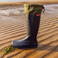 A single black Pokeboo TREAD rubber rain boot with an olive green cuff stands on wet sand at the edge of the sea, with rippled sand, water, and a distant shoreline visible in the background.